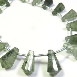 May include: A strand of faceted, trapezoidal green rutilated quartz beads arranged in a circular pattern. Each translucent stone features dark, needle-like inclusions and is strung on a thin, silver-coloured metal wire, creating a uniform sequence.