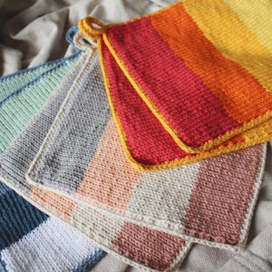 Könnte beinhalten: Ein Set aus sechs gestrickten Topflappen in verschiedenen Farben, darunter Grün, Grau, Pfirsich und Rot. Die Topflappen sind gestreift und haben einen gehäkelten Rand.