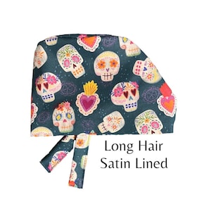 May include: A teal blue surgical scrub hat with a colorful sugar skull pattern. The hat has long ties and is lined with satin. The text "Long Hair Satin Lined" is printed on the hat.