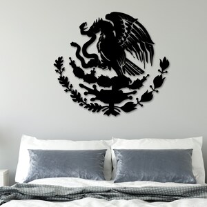 Op de afbeelding: Zwarte metalen wandkunst die het Mexicaanse wapenschild afbeeldt, met een adelaar die op een cactus zit met een slang in zijn bek. De adelaar wordt omringd door een krans van bladeren en bloemen.