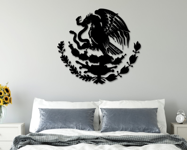 Op de afbeelding: Zwarte metalen wandkunst die het Mexicaanse wapenschild afbeeldt, met een adelaar die op een cactus zit met een slang in zijn bek. De adelaar wordt omringd door een krans van bladeren en bloemen.