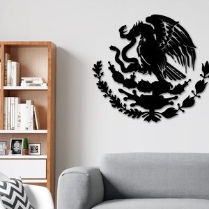 Op de afbeelding: Zwarte metalen wandkunst die het Mexicaanse wapenschild afbeeldt, met een adelaar die op een cactus zit met een slang in zijn bek. De adelaar wordt omringd door een krans van bladeren.