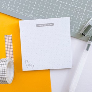 May include: A white notepad with the text "BRAIN DOODLES" and a small dinosaur illustration, a roll of black and white grid tape, and a clear pen with a gray cap are arranged on a yellow and gray surface.