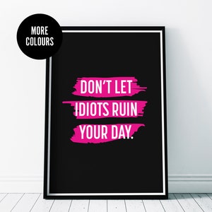以下が含まれることがあります： 黒いフレームに入ったアートプリント。テキスト「DON'T LET IDIOTS RUIN YOUR DAY」は白とピンクの筆致。左上隅には「MORE COLOURS」という文字が入った黒い円があります。