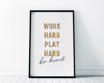 May include: A framed white letter board with the words "WORK HARD PLAY HARD be kind" in gold and black lettering. The board has a black frame and is displayed against a white wall.
