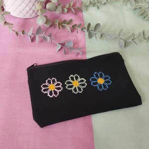 Puede incluir: Bolsa negra con cremallera y tres flores de margarita bordadas en rosa, verde y azul, cada una con un centro amarillo. La bolsa es rectangular y parece estar hecha de una tela suave. El fondo es una mezcla de rosa y verde.