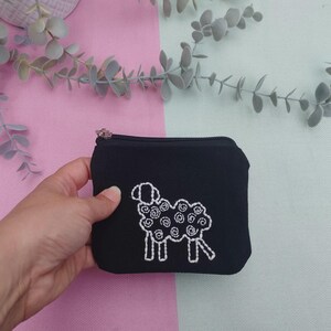 Puede incluir: Pequeño monedero negro con cremallera y un diseño de oveja bordada en blanco. La oveja tiene un patrón de lana rizada. El monedero está sobre un fondo rosa y verde menta. Una mano sostiene el monedero.