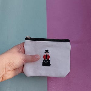 Puede incluir: Un pequeño estuche de lona blanca con cremallera que presenta un diseño de punto de cruz de una figura con sombrero de copa negro, chaqueta roja y falda negra. El estuche tiene un cierre de cremallera negro en el borde superior.