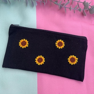Puede incluir: Un estuche rectangular negro con cremallera que presenta cuatro motivos de girasoles bordados. Cada girasol tiene un centro marrón oscuro rodeado de pétalos de color amarillo brillante. El estuche está hecho de tela texturizada y cierre negro.