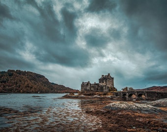 May include: A gray stone castle with a bridge leading to it, set against a dramatic cloudy sky. The castle is situated on a small island in a body of water.