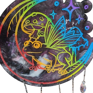 May include: A decorative wind chime with a frog and mushroom design, set against a galaxy-themed crescent moon. The artwork features rainbow colours and dangling crystal beads. A charming piece for a garden or home, with a celestial theme.