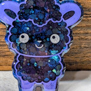May include: A whimsical, handcrafted sheep-shaped resin charm. The charm features a vibrant mix of dark blue, purple, and teal glitter, creating a galaxy-like effect. The sheep has large, silver-colored eyes and a smiling mouth, with a light purple outline.