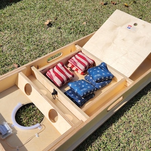 May include: A wooden cornhole game set with a natural finish, featuring red and white striped bean bags and blue bean bags with white stars. The set includes a hinged top and a circular target hole.