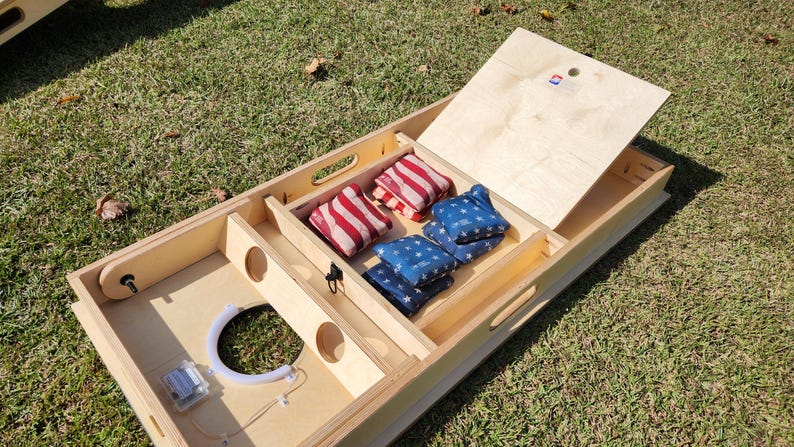 May include: A wooden cornhole game set with a natural finish, featuring red and white striped bean bags and blue bean bags with white stars. The set includes a hinged top and a circular target hole.