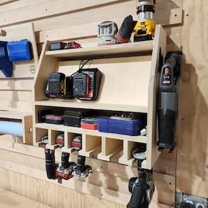 Puede incluir: Un organizador de pared de madera para herramientas el&eacute;ctricas. Cuenta con estantes para guardar bater&iacute;as y un estante para colgar herramientas. El organizador est&aacute; hecho de madera clara y tiene un acabado natural. Hay varias herramientas el&eacute;ctricas colgadas en el estante, incluyendo una sierra, un taladro y una lijadora.