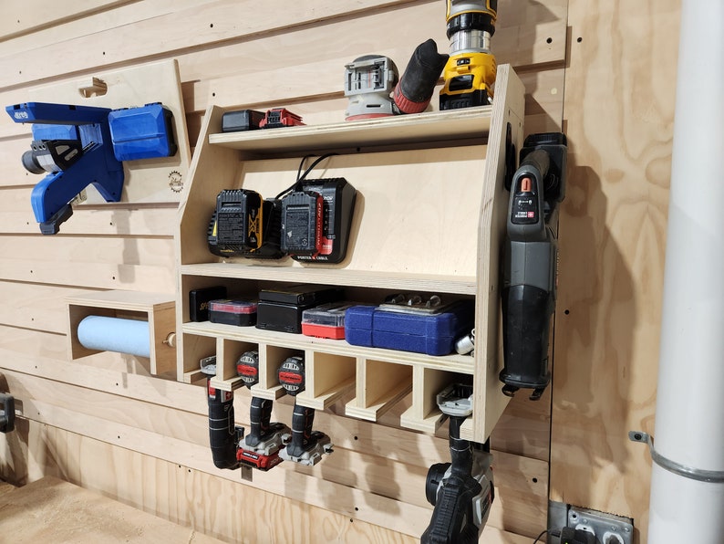 Puede incluir: Un organizador de pared de madera para herramientas el&eacute;ctricas. Cuenta con estantes para guardar bater&iacute;as y un estante para colgar herramientas. El organizador est&aacute; hecho de madera clara y tiene un acabado natural. Hay varias herramientas el&eacute;ctricas colgadas en el estante, incluyendo una sierra, un taladro y una lijadora.