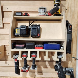 Puede incluir: Un estante de almacenamiento de madera montado en la pared con estantes para herramientas el&eacute;ctricas y bater&iacute;as. El estante tiene ranuras para sujetar taladros y un estante para guardar bater&iacute;as de herramientas. El estante est&aacute; hecho de madera clara y tiene un acabado natural.