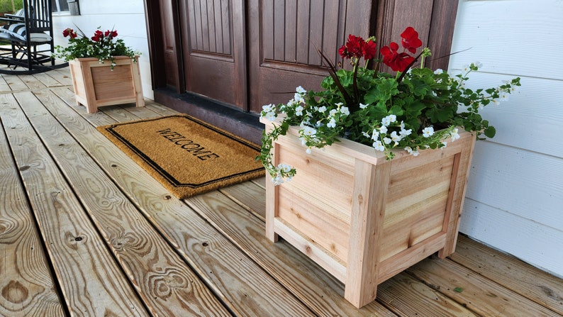 Puede incluir: Dos macetas de madera con flores rojas y blancas en un porche de madera con una alfombra de bienvenida.