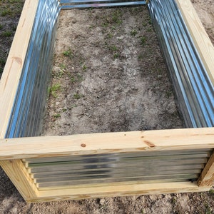 May include: Rectangular raised garden bed with corrugated metal sides and a natural wood frame. The bed is empty and sits on a patch of dirt and grass, ready for planting. Another similar bed is visible in the background.