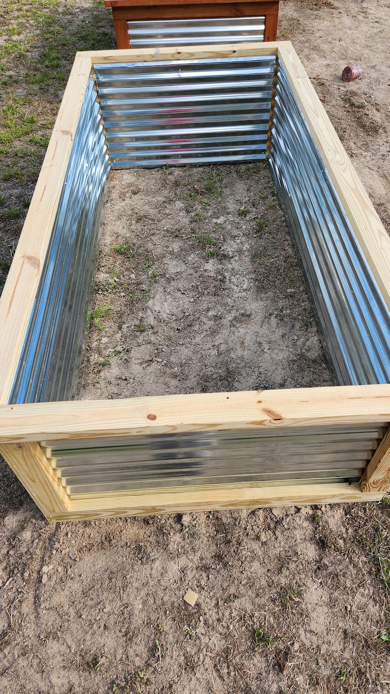 May include: Rectangular raised garden bed with corrugated metal sides and a natural wood frame. The bed is empty and sits on a patch of dirt and grass, ready for planting. Another similar bed is visible in the background.