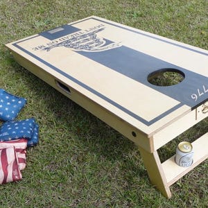 May include: A wooden cornhole board with a cream-colored surface and a dark blue stripe down the center. The board has a hole for tossing bean bags, and the words "DON'T TREAD ON ME" are visible. Several red, white, and blue bean bags are scattered around.