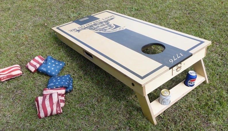 May include: A wooden cornhole board with a cream-colored surface and a dark blue stripe down the center. The board has a hole for tossing bean bags, and the words "DON'T TREAD ON ME" are visible. Several red, white, and blue bean bags are scattered around.