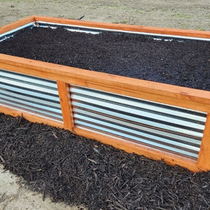 May include: Two rectangular raised garden beds with corrugated metal sides and wooden frames. The beds are filled with dark soil and sit on a bed of dark mulch. The wooden frames are a warm, natural color.