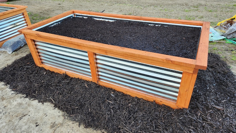 May include: Two rectangular raised garden beds with corrugated metal sides and wooden frames. The beds are filled with dark soil and sit on a bed of dark mulch. The wooden frames are a warm, natural color.