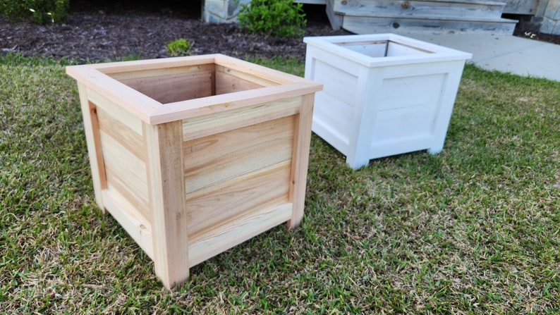 Puede incluir: Dos jardineras cuadradas de madera, una de madera natural y otra pintada de blanco, sentadas en un c&eacute;sped verde.