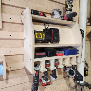 Puede incluir: Una unidad de almacenamiento de madera montada en la pared con estantes para herramientas el&eacute;ctricas y bater&iacute;as. La unidad tiene un cable de alimentaci&oacute;n negro y contiene una variedad de herramientas, incluyendo una lijadora, una fresadora y varios taladros. La unidad est&aacute; montada en una pared de madera clara.