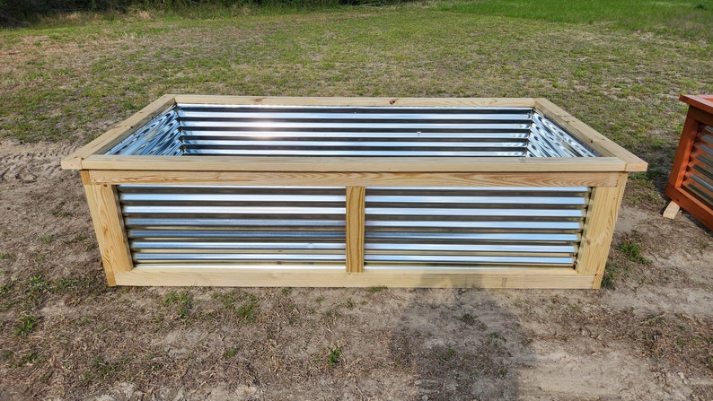 May include: A rectangular raised garden bed with a wooden frame and corrugated metal sides. The natural wood frame surrounds the galvanized metal, creating a rustic look. The bed is designed for planting flowers, vegetables, or herbs.