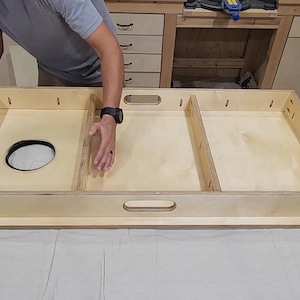 May include: A light-colored wooden cornhole board frame with a circular hole and two rectangular compartments. The frame has a handle cut out on the side. The board is being assembled on a white surface.