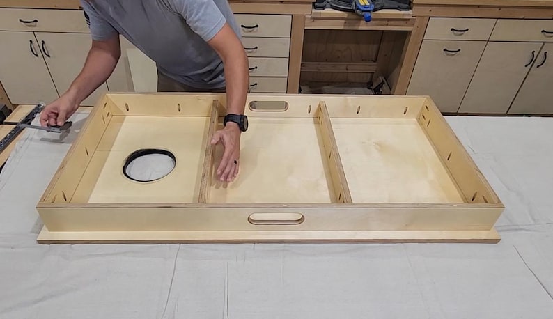 May include: A light-colored wooden cornhole board frame with a circular hole and two rectangular compartments. The frame has a handle cut out on the side. The board is being assembled on a white surface.