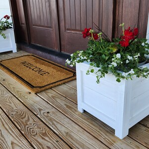 Puede incluir: Dos macetas de madera blancas con flores rojas y blancas en un porche de madera con una alfombra de bienvenida.
