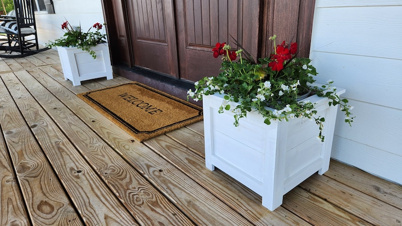 Puede incluir: Dos macetas de madera blancas con flores rojas y blancas en un porche de madera con una alfombra de bienvenida.