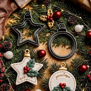 May include: Two dark grey plastic cookie cutters in star and circular ornament shapes, alongside two matching white cookie decorations with gold caps, green holly leaves, red berries, and the names ava and penelope written in brown script.