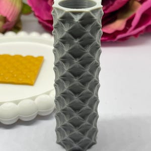 May include: A grey, cylindrical vase with a textured, diamond-like pattern. The vase has a hexagonal opening at the top. The background includes a white plate with a yellow object and pink flowers.