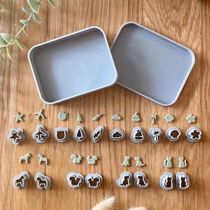 May include: A collection of small, light green clay shapes and matching grey plastic cutters arranged in rows. The shapes include stars, aeroplanes, tulips, trees, lightning bolts, clouds, sailboats, palm trees, shells, flowers, horses, squirrels, dinosaurs, and cats.