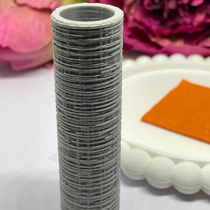 May include: A cylindrical, gray object with a woven pattern, possibly a hair curler. The top edge is white. An orange square and a white plate with a scalloped edge are in the background, along with pink flowers.
