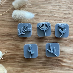 May include: Five square, slate blue stamps featuring raised botanical designs, including a sprig with berries, a fan-shaped flower, and three different leaf patterns. Each stamp has rounded corners and a smooth, matte finish on the surface.