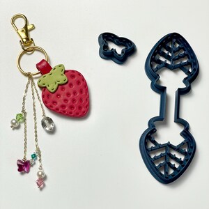 May include: A red strawberry keychain with a gold clasp and dangling charms. Next to it are two dark blue cookie cutters: a butterfly and a leaf-shaped design. The strawberry has a green leaf and a textured surface.