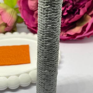 May include: A grey, cylindrical candle with a woven texture. The candle has a white top and is set against a backdrop of pink flowers and a white plate with an orange square.