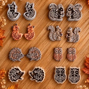 May include: A collection of eight pairs of animal-shaped earrings featuring foxes, squirrels, hedgehogs, and owls. Each pair displays intricate floral and leaf patterns embossed on the surface, rendered in various shades of brown, grey, and cream.