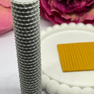 May include: A grey, cylindrical candle with a textured, honeycomb-like pattern. The candle stands on a white plate with a yellow, textured square. Pink and purple flowers are in the background.