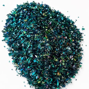 May include: A collection of small, irregular crushed shell fragments in deep shades of teal, emerald green, and dark blue. The pieces have a reflective, iridescent finish that catches the light, creating a shimmering effect across the pile.
