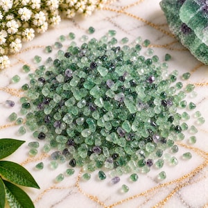 May include: A large collection of small, tumbled fluorite gemstones in shades of translucent light green and deep purple. The stones are irregular in shape and size, featuring a polished, glossy finish that reflects light across the scattered pile.