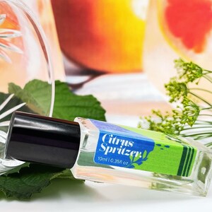 May include: A clear glass bottle with a black cap labelled "Citrus Spritzer" with blue and green accents. The bottle is lying on a green leaf, with a crystal glass and a blurred background.