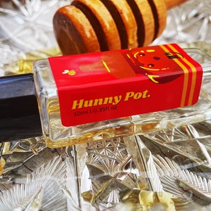 May include: A small, clear glass bottle with a black cap and a red and yellow label that says "Hunny Pot. 10ml | 0.35fl oz".