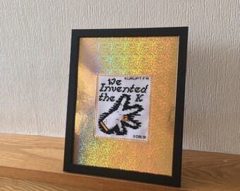 May include: A framed cross-stitch with a gold glitter background. The cross-stitch features a black and white illustration of a hand with the text "We Invented the K" and "KURUPT FM 108.9".