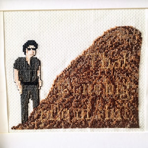 May include: Cross-stitch artwork depicting a man in sunglasses and a dark shirt standing on a brown hill. The hill is made of cross-stitched brown and beige squares.